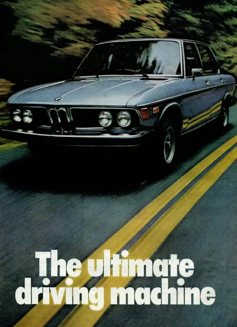 Vintage auto ad: The Ultimate Driving Machine A vintage light blue BMW sedan, seen from the front three-quarter angle, driving on a dark asphalt road with two bright yellow lines. The background is a blurry wash of green and brown foliage, suggesting motion. White text at the bottom reads 'The ultimate driving machine'.